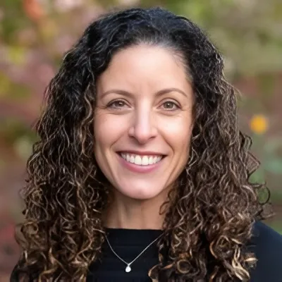 Natural Pharmaceuticals OmegaMarine™ Premium 
                                Uśmiechnięta kobieta o ciemnych, kręconych włosach, 
                                w czarnym topie z delikatnym naszyjnikiem; portret na zewnątrz, 
                                rozmyte tło zieleni i jesiennych kolorów.
                            