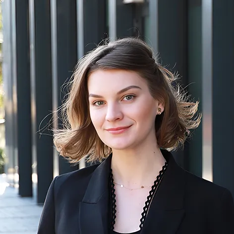 Natural Pharmaceuticals OmegaMarine™ Premium Portret uśmiechniętej kobiety w czarnym żakiecie, kadr do ramion, na tle nowoczesnego przeszklonego budynku.