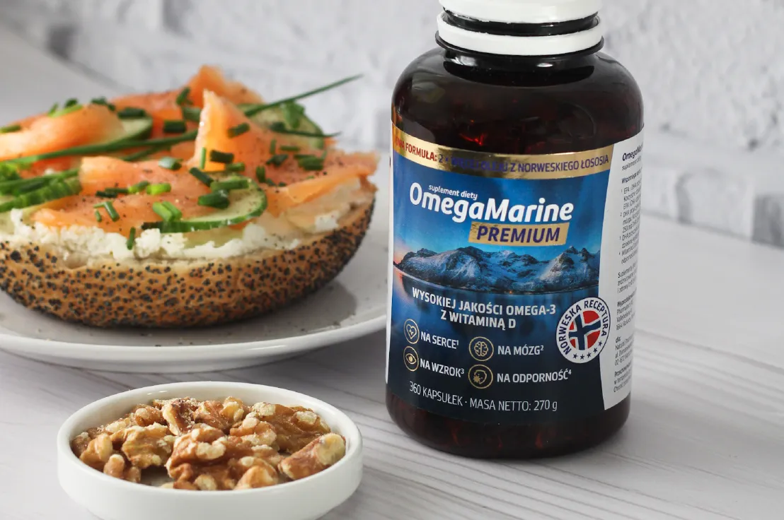 Natural Pharmaceuticals OmegaMarine™ Premium Butelka suplementu OmegaMarine™ Premium (omega‑3 z witaminą D) na blacie; obok bajgiel z łososiem i ogórkiem oraz miseczka orzechów włoskich.