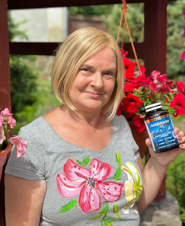 Kobieta w ogrodzie trzyma butelkę suplementu OmegaMarine™ Premium; w tle czerwone kwiaty, na koszulce motyw kwiatowy.