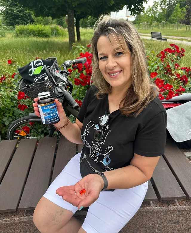 Kobieta siedząca na ławce przy rowerze i czerwonych kwiatach; uśmiecha się, trzyma butelkę OmegaMarine™ Premium i kilka czerwonych kapsułek na dłoni.