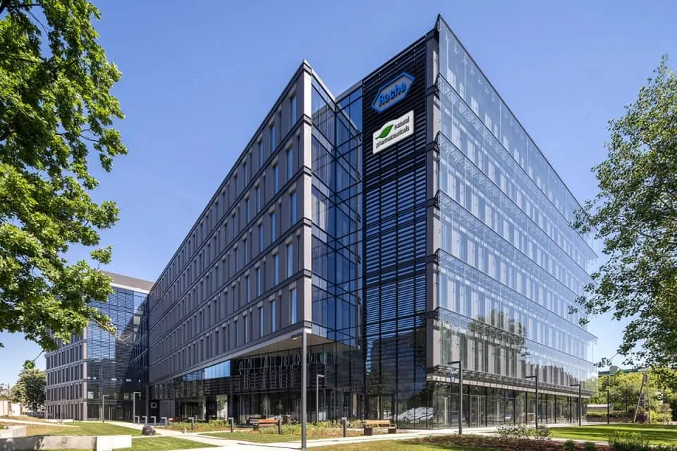 Natural Pharmaceuticals OmegaMarine™ Premium Nowoczesny przeszklony biurowiec w słoneczny dzień; elewacja z logotypami i otoczenie z zielenią oraz alejkami.