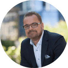 Natural Pharmaceuticals OmegaMarine™ Premium Portret uśmiechniętego mężczyzny w okularach i marynarce na rozmytym miejskim tle.